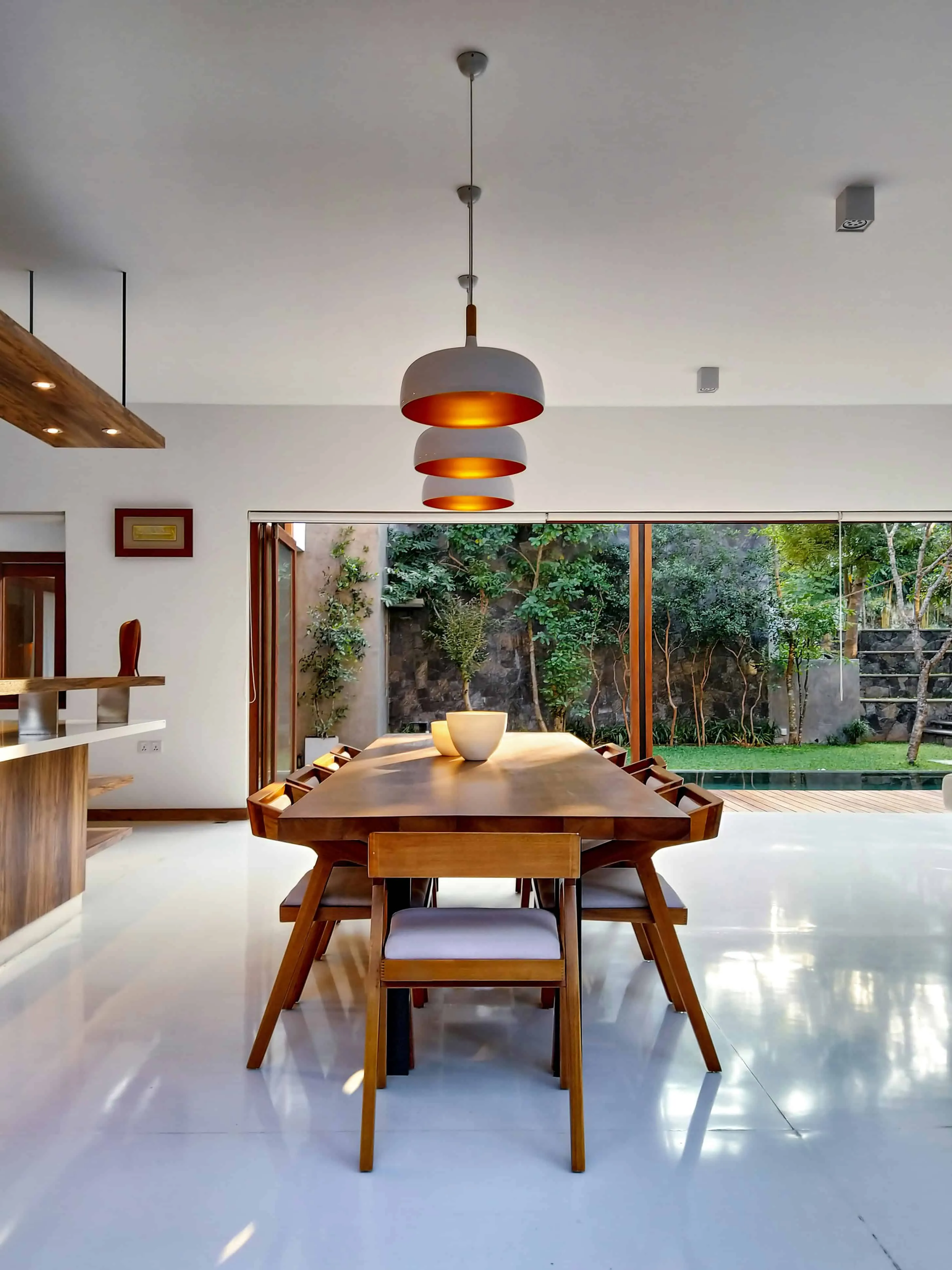 Elegant wooden dining table with modern pendant lights in a bright open-concept kitchen and outdoor garden area.